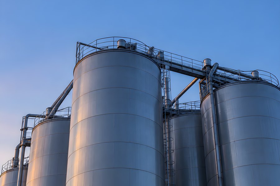 Tall metal grain silos linked by a rooftop catwalk against a blue evening sky