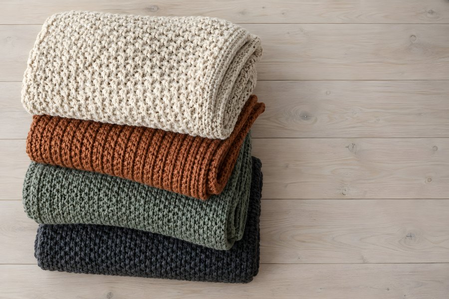 Stack of folded chunky knit wool blankets in cream, rust, sage and navy on a wooden floor with copy space