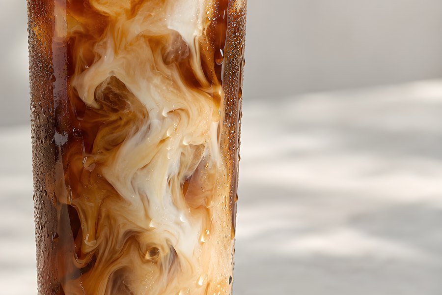 Iced latte with milk swirling into dark coffee in a tall glass, ice cubes and condensation on a sunlit concrete surface