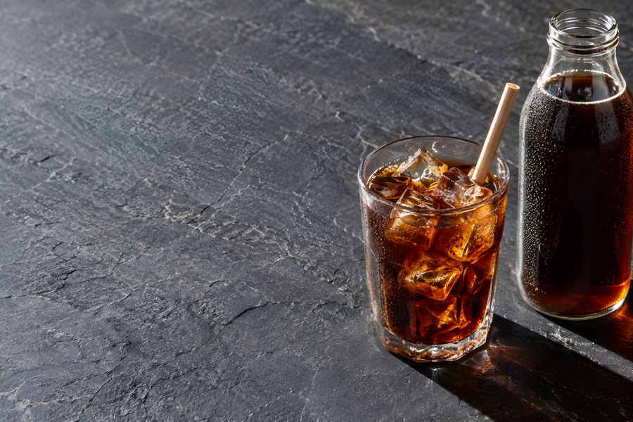 Iced cold brew coffee in a glass tumbler beside a filled glass bottle on dark slate with condensation and dramatic side light