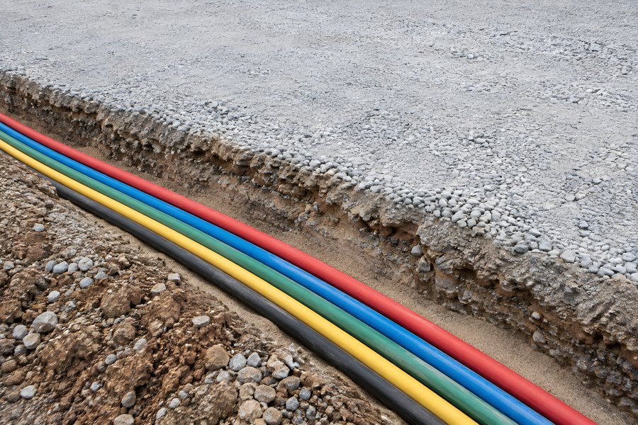 Five color-coded conduit pipes in an open trench beside a cut gravel road base for underground cable installation