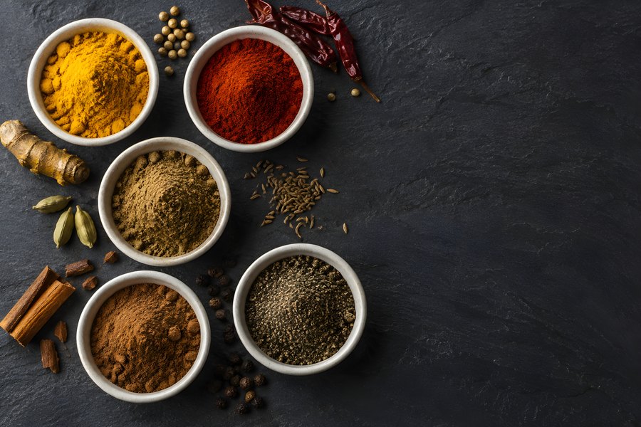 Ground spices in white bowls with whole chili peppers, cinnamon sticks, cardamom pods and ginger root on a dark slate background, overhead flat lay