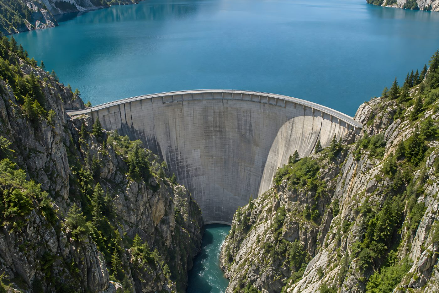 Aerial view of a curved concrete arch dam holding back a turquoise mountain reservoir above a narrow rocky gorge