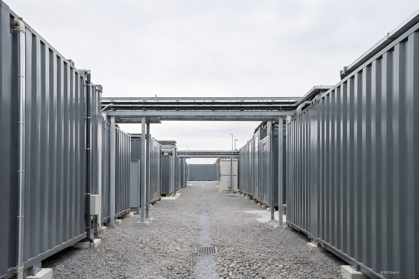 Battery energy storage system site with grey steel containers and an overhead pipe bridge along a gravel service aisle under overcast sky