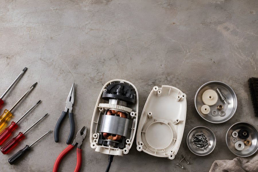 Disassembled small appliance motor with screwdrivers, pliers, gears and spare parts on a repair workbench, top view flat lay