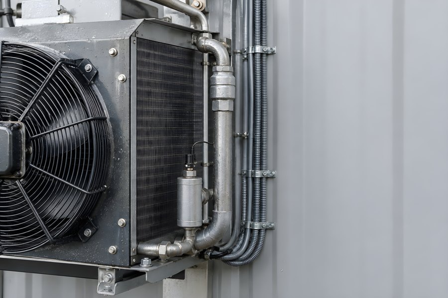 Close-up of a fan-cooled heat exchanger with steel pipework and cable conduits on a grey battery energy storage container