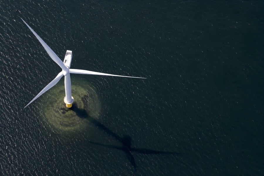 Aerial view of a single offshore wind turbine casting a long shadow on dark rippled sea water around its yellow monopile base
