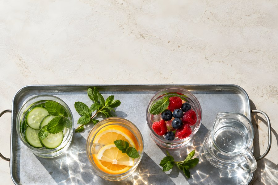 Overhead view of fruit-infused water glasses with cucumber, citrus and berries beside a water pitcher on a sunlit tray