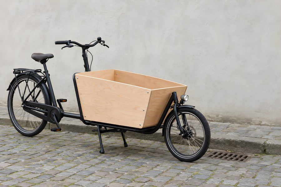 Front-loading cargo bike with an empty plywood box parked beside a stone wall