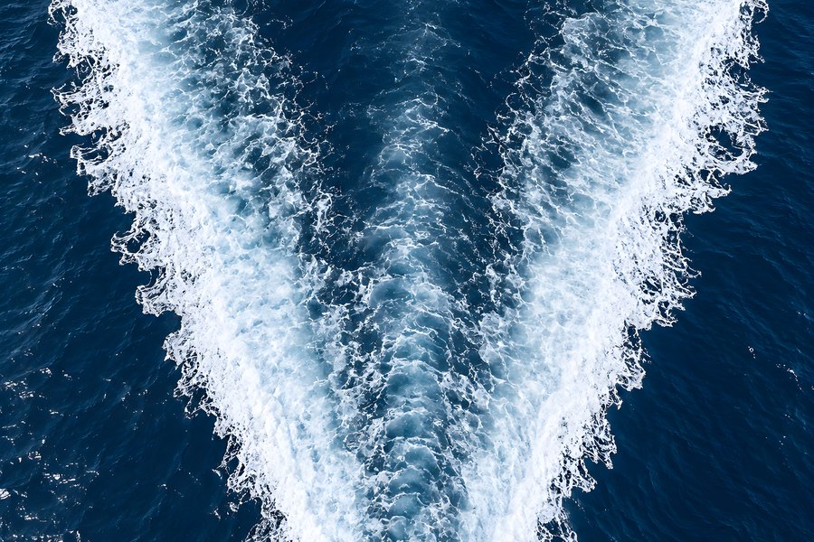 Aerial top-down view of a white V-shaped boat wake churning across deep blue open ocean water, ferry wake trail with no vessel or land visible