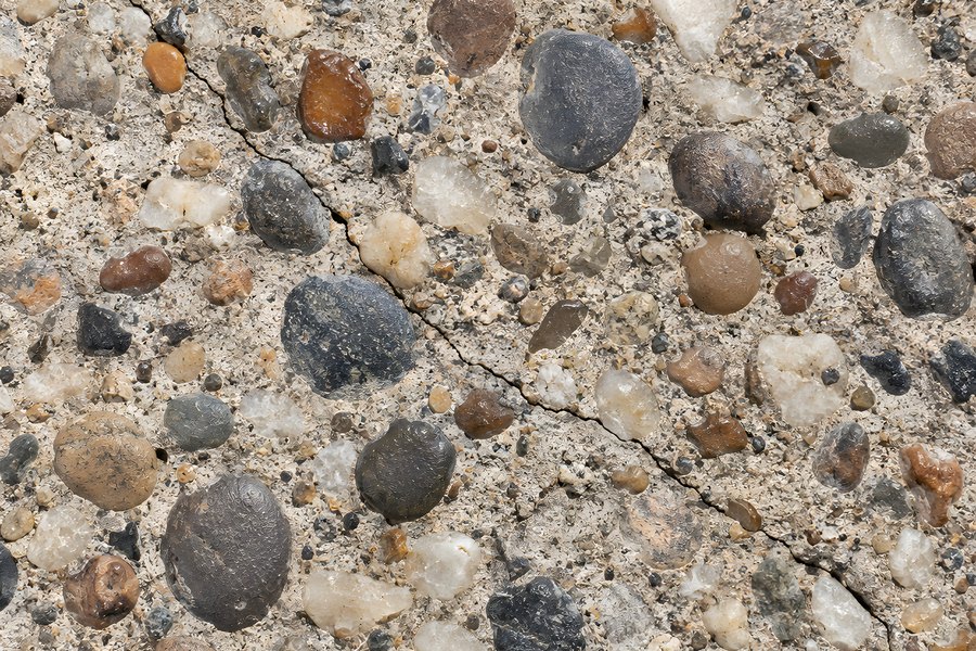 Close-up of exposed aggregate concrete texture with round pebbles, gravel, and a hairline crack across a rough grey-beige surface background