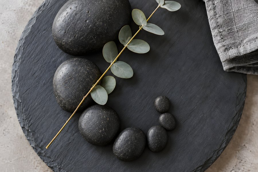 Black basalt hot stones arranged in a curved row with a eucalyptus sprig and folded linen towel on a round slate tray, overhead flat lay